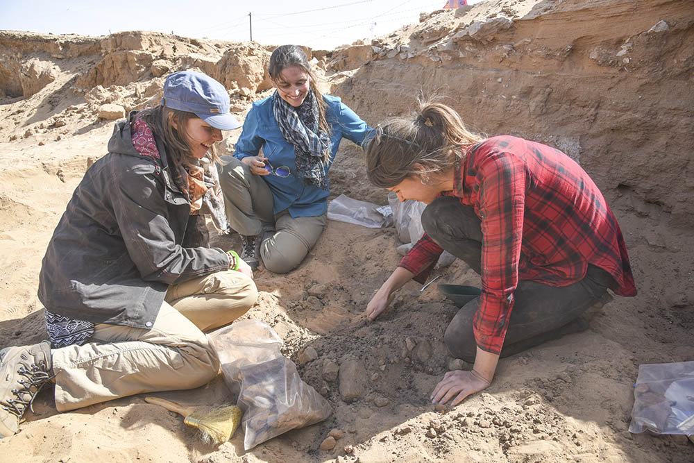 Three seated figures excavating objects in the mud brick building.
