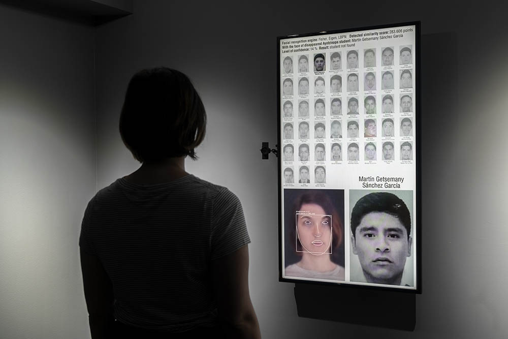 Figure standing in front of a vertical video screen depicting a facial-recognition system.