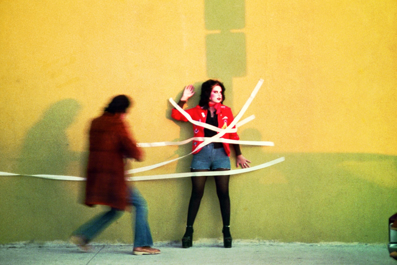 Asco artist Patssi Valdez is being taped to a yellow wall in East LA by fellow Asco member Gronk. Patssi has a full face of makeup and wears a red jacket short shorts with tights and platform heels.