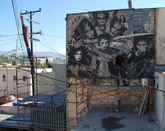 Willie Herron III paints a mural on a Los Angeles roof while standing on scaffolding. The background shows multiple other buildings and mountains.