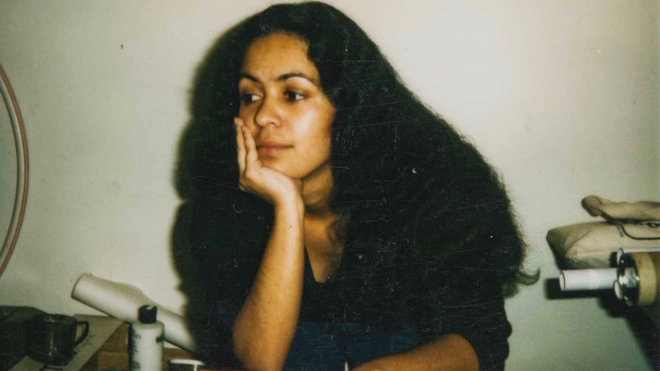 Frances Carrasco sits with her head resting in her right hand. While posing she gazes to the left at something off camera.