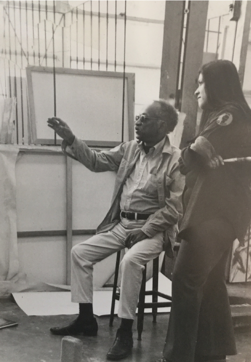 Charles White and Judithe Hernandez talk in a classroom with a large amount of natural light. Judithe holds a paintbrush and White is pointing to something out of frame suggesting he is providing feedback on her work.