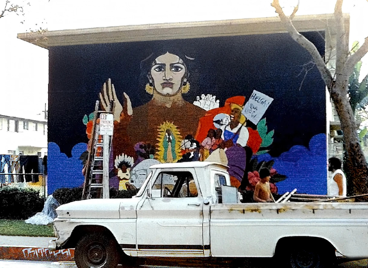 A pickup truck with supplies is parked in front of the site for the mural by Judithe Hernandez. The mural is not complete and the original brown building color is visible.