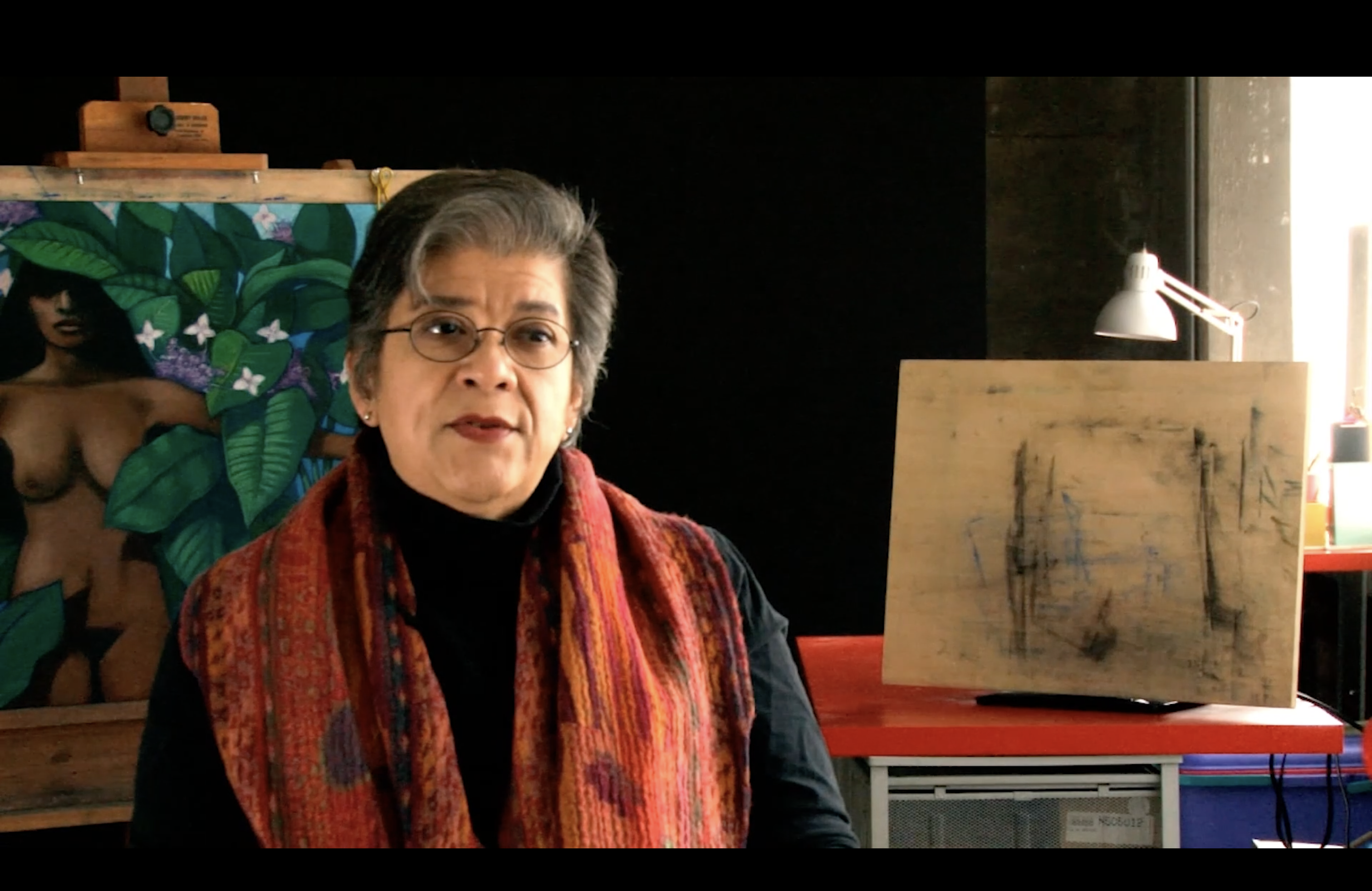 Judithe Hernandez is center frame being interviewed. A canvas of a woman surrounding the fauna and an easel without a work on it are in the background.