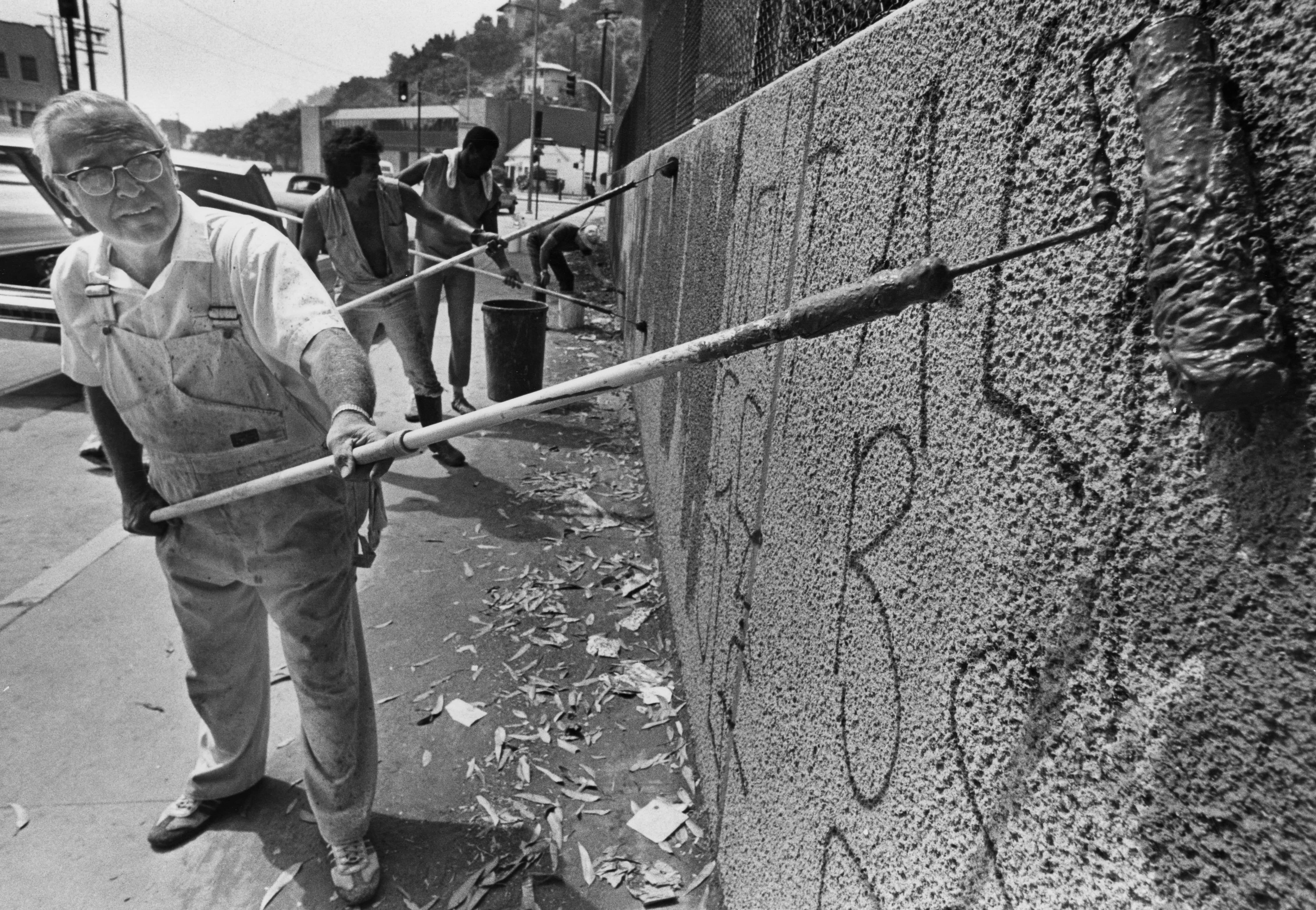 A group of four men hold long paint rollers as they cover up a short call with graffiti writing. The paint they are using is darker than the wall displaying the patches where tags were painted. In the extreme foreground a paint roller soaked with paint is deliberately prominent.