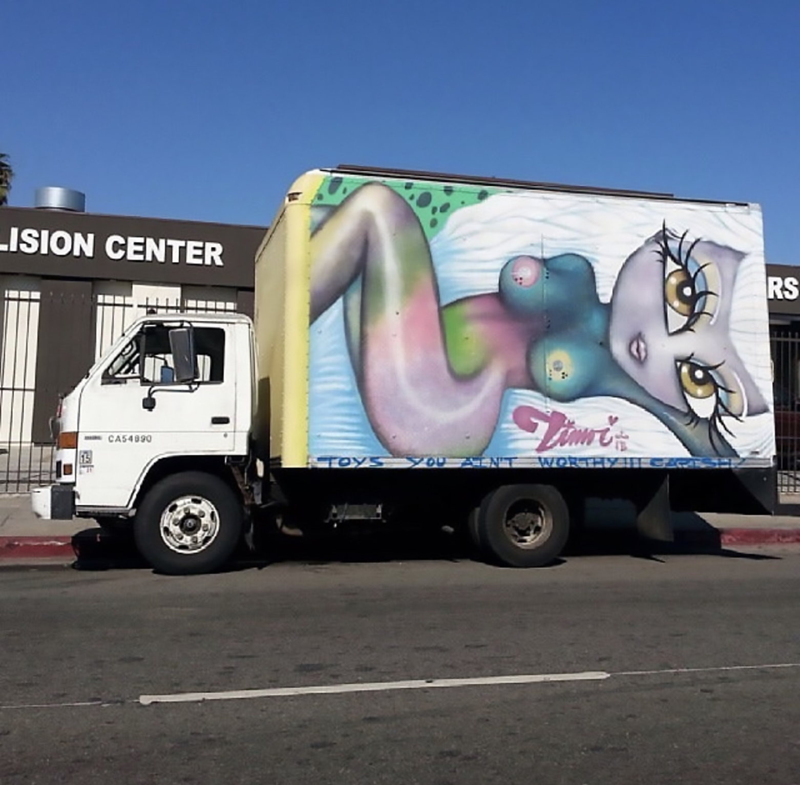 A box truck sits on the street with a Timoi spray painted work featuring a nude femme character in blue purple and green. The figure has exaggerated eyes and there is text below them reading toys you aint worthy with exclamation points.