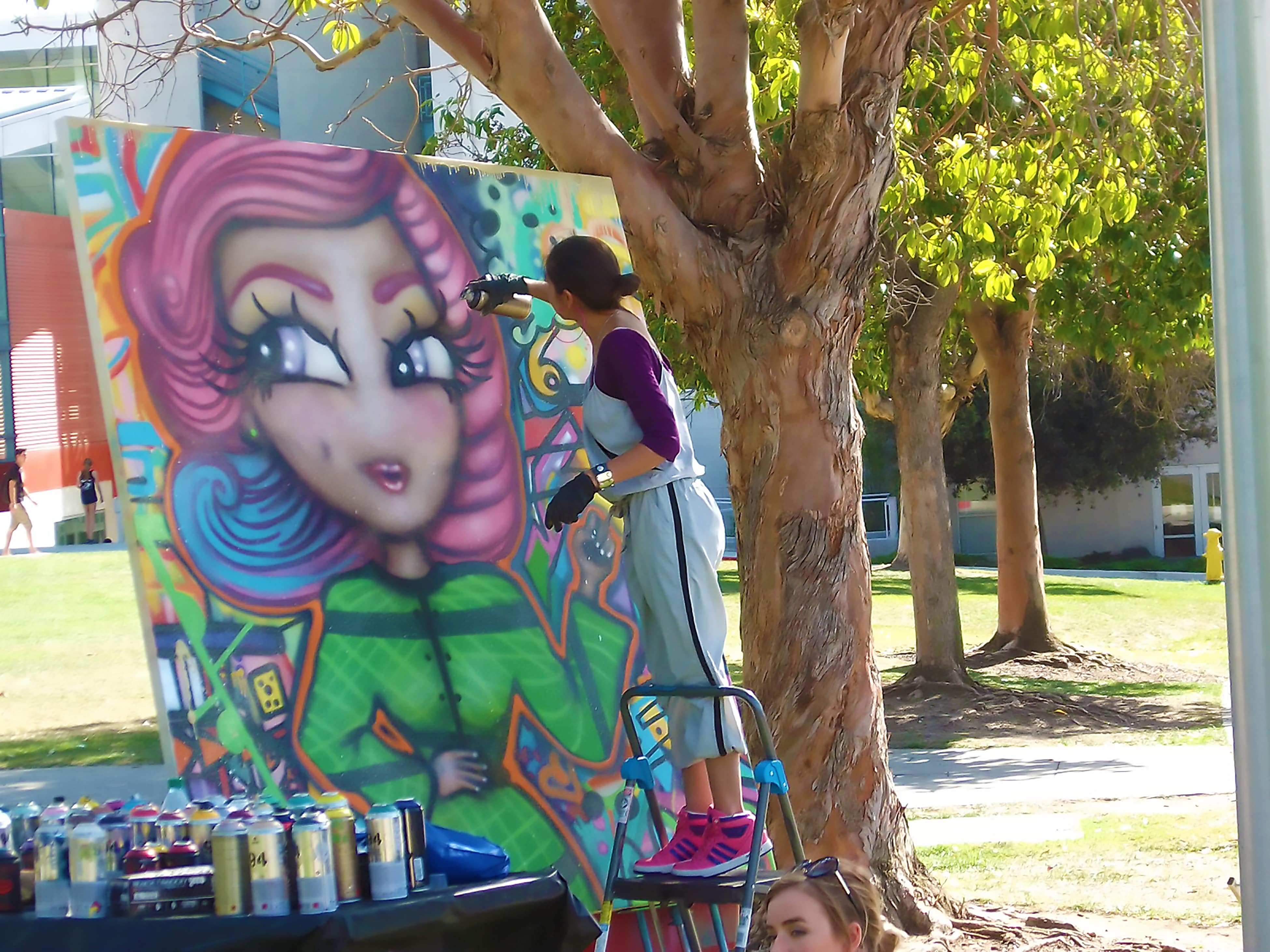 Timoi spray paints a portrait of Luisa Moreno who appears in many colors including pink green and blue. In front of the canvas rests a large table filled with spray cans of various colors.