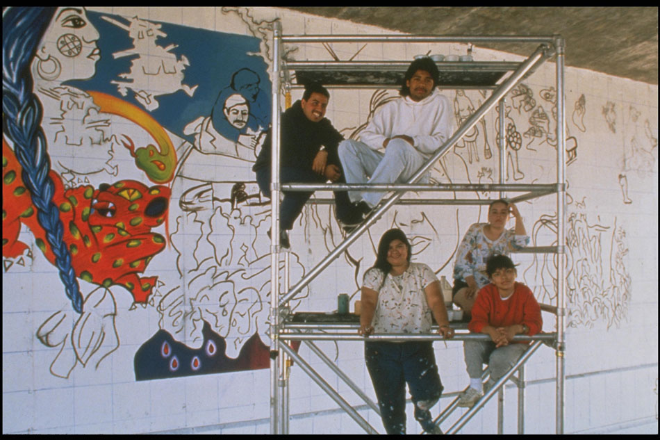 A group of five individuals including Yreina Cervantez rest on scaffolding in front of the early stages of the mural La Ofrenda. The grids utilized to paint the mural in a coordinated manner are visible.