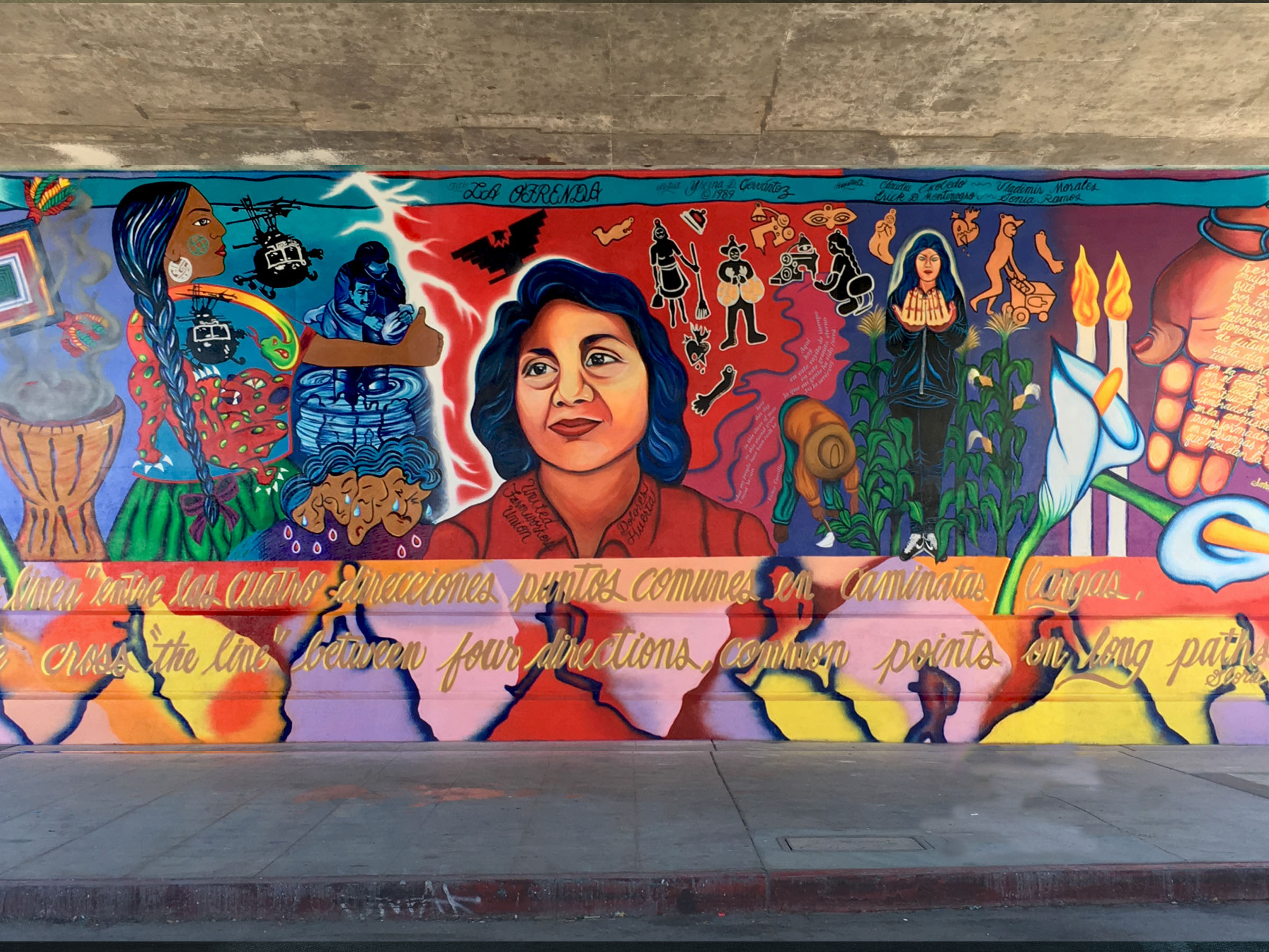 The complete mural underneath a freeway overpass with an unhoused encampment set
                            up in front of the Yreina Cervantez mural.