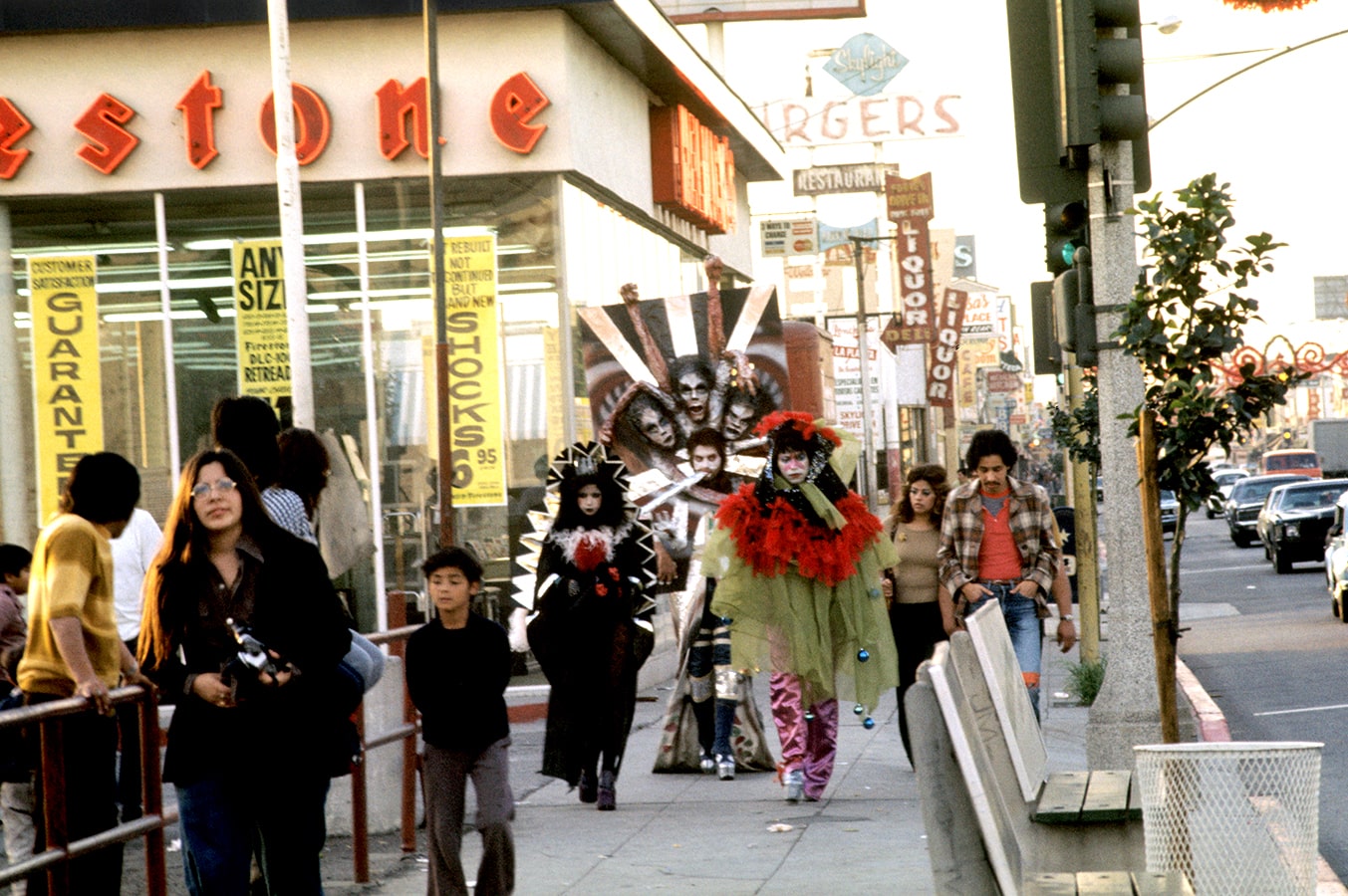 A trio of artists (Asco) dressed in costume walks down Whittier Boulevard surrounded by a
                            local crowd.