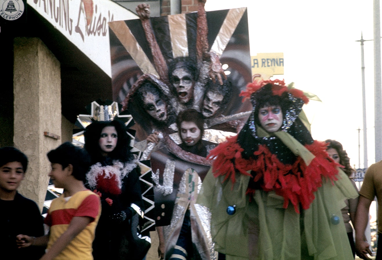 A trio of artists dressed in costume walks down Whittier Boulevard surrounded by a local crowd (second image).