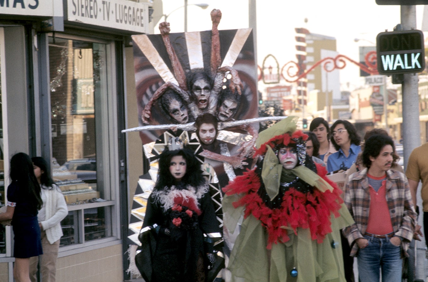 A trio of artists dressed in costume walks down Whittier Boulevard surrounded by a local crowd (third image).