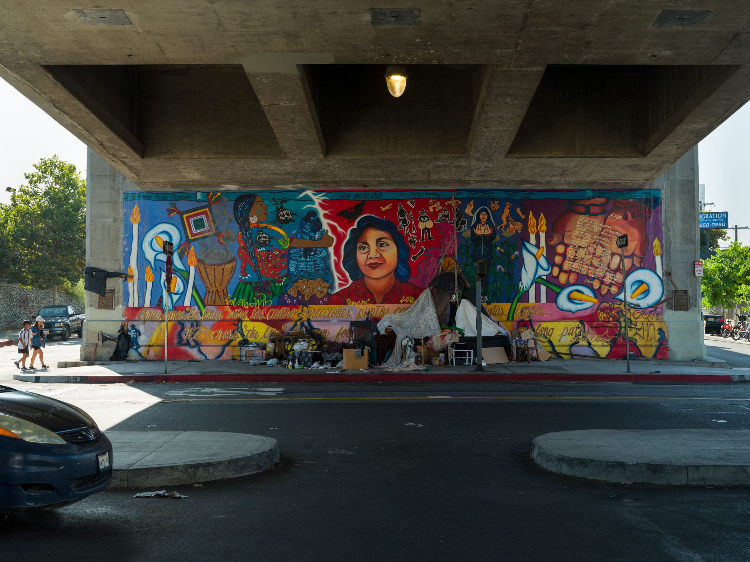 The complete mural underneath a freeway overpass with an unhoused encampment set up in front of the Yreina Cervantez mural.