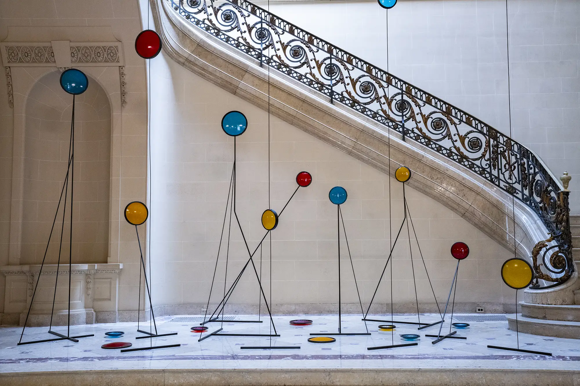 
A modern art sculpture featuring colorful, glass-like discs hangs from the ceiling and stands on the floor in a grand, ornate marble entryway with a large staircase. The discs are in various sizes and colors, including red, yellow, and blue. They are suspended by thin, black wires that also hold up some of the discs from the floor. The staircase has a decorative, black wrought-iron railing with gold accents.