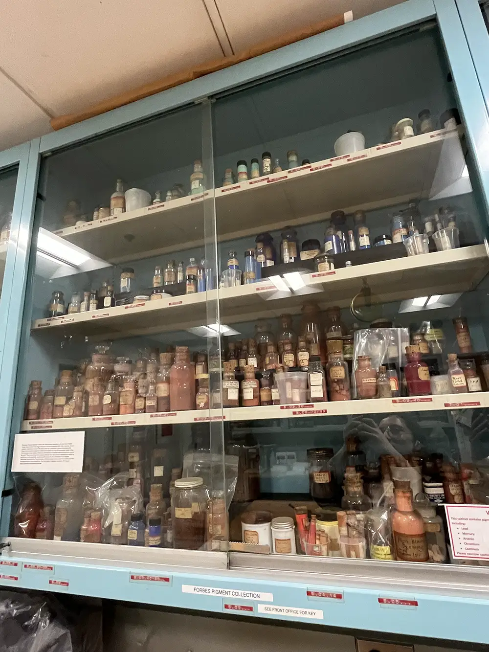 
A tall, glass-fronted cabinet filled with dozens of small, vintage bottles and jars, containing various powders, liquids, and pigments. The cabinet is organized with several shelves, and many of the containers have old, handwritten labels. A sign on the bottom shelf says 