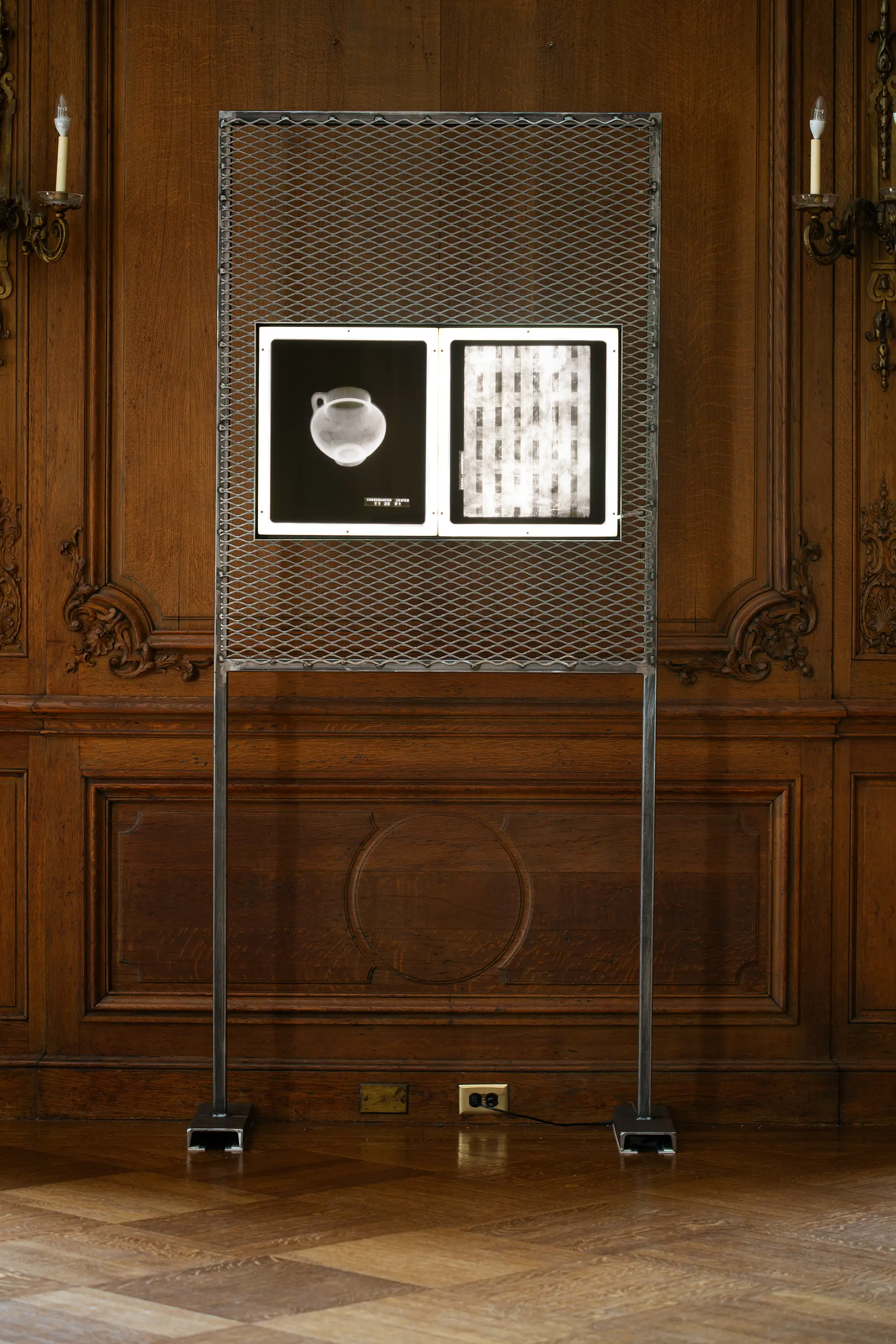 
A tall, industrial-style metal stand with a mesh frame holds two backlit images. The image on the left is a white X-ray of an ancient-looking ceramic vessel. The image on the right is a blurred, black-and-white X-ray of what looks like a set of keys or small objects. The stand is positioned in the center of a room with ornate wooden paneling and a parquet floor.