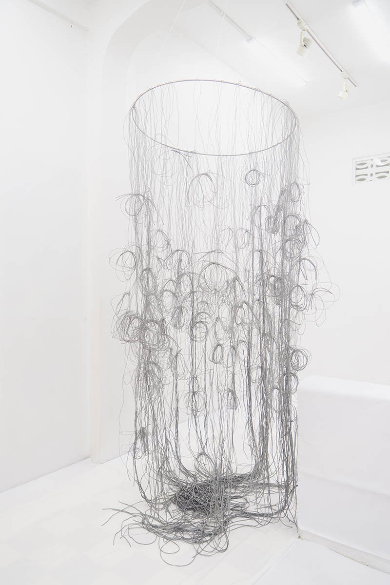 Suspended cylindrical sculpture of tangled silver wire hanging from a circular frame in a white gallery.