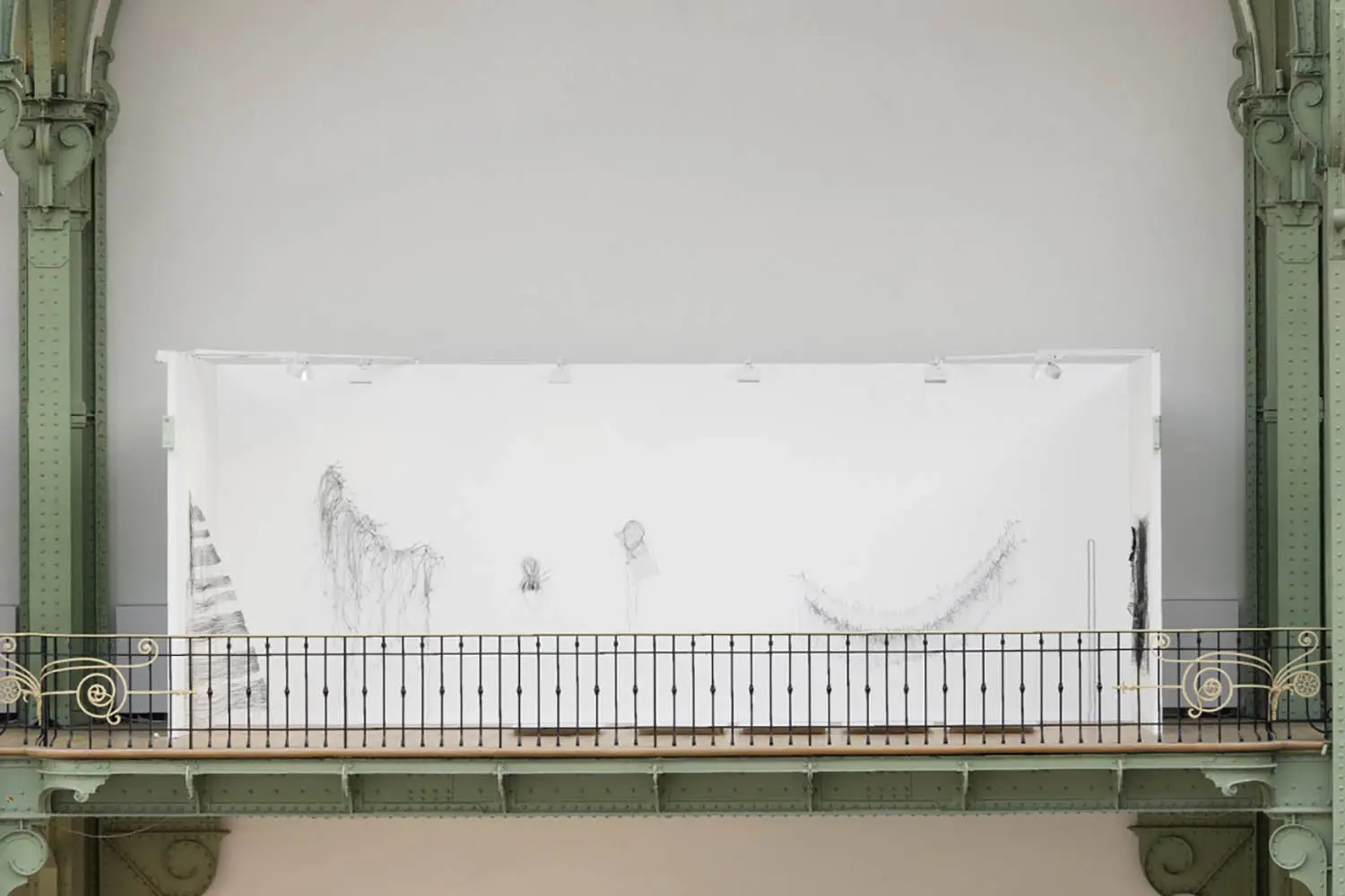 Balcony view framed by ornate green architectural ironwork looking onto a white exhibition wall with delicate wire sculptures.