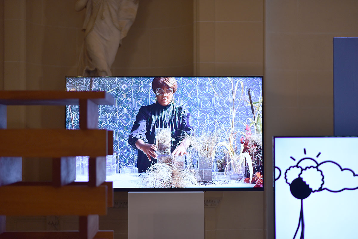 Installation view: Two large flat screen monitors, out of focus scultpure in foreground.