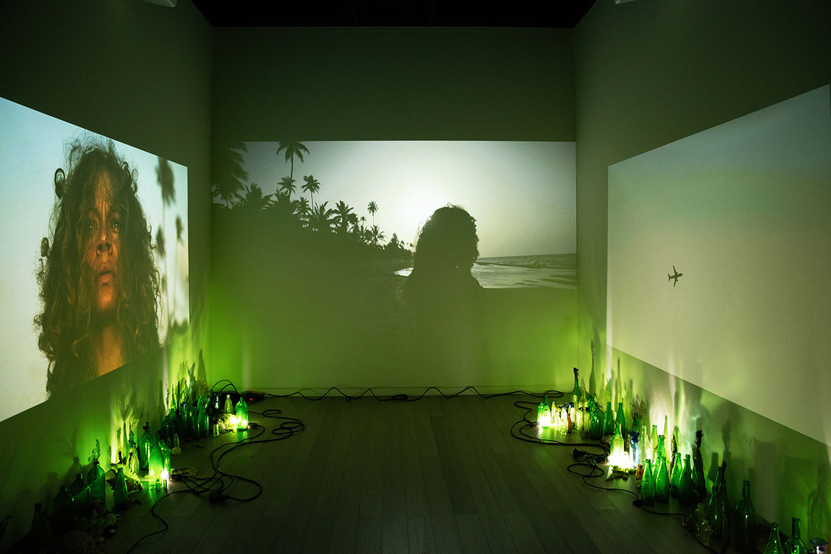 Three video projections of a woman on a tropical island from different prospectives. The room is illuminated with light emanating from green glass bottles displayed on the floor.